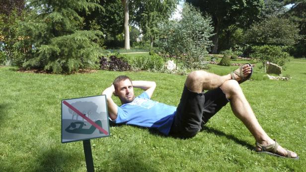 Ein Mann liegt auf einer Wiese neben einem Schild, das das Liegen verbietet.