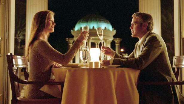 George Clooney und eine Frau stoßen in einem eleganten Restaurant mit Champagner an.