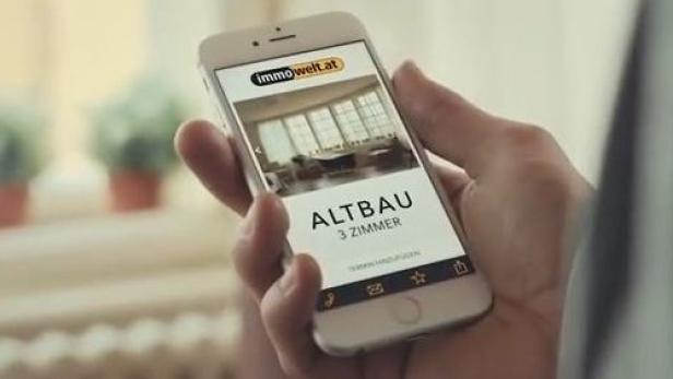 Eine Hand hält ein Smartphone mit der Webseite immowelt.at, die eine 3-Zimmer-Altbauwohnung zeigt.