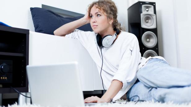Eine Frau sitzt mit Kopfhörern auf dem Teppich und benutzt einen Laptop.