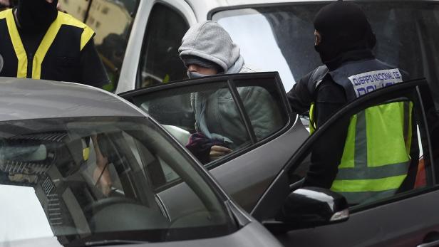 Eine Person mit Kapuze steigt in ein Auto, bewacht von zwei Polizisten mit Sturmhauben.