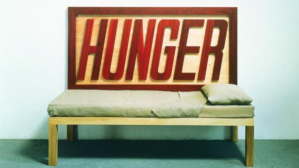 Ein Bett steht vor einer Wand, über dem Bett hängt ein Schild mit dem Wort „Hunger“.