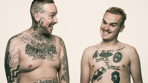 Zwei Männer mit Tätowierungen auf ihren Oberkörpern stehen sich gegenüber und lächeln.