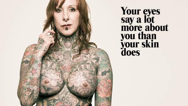 Eine Frau mit zahlreichen Tätowierungen und Piercings posiert vor einem hellen Hintergrund.