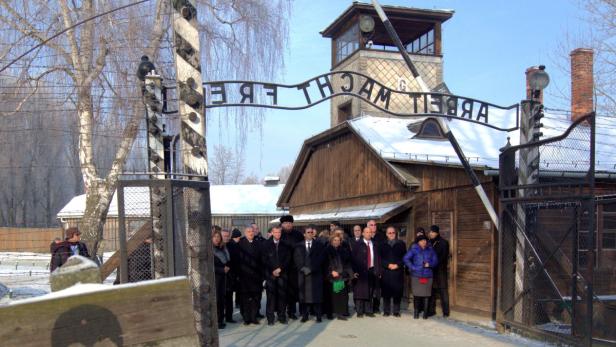 Eine Gruppe von Menschen steht vor dem Eingangstor mit der Aufschrift „Arbeit macht frei“ in Auschwitz.