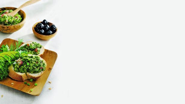 Zwei Brotscheiben belegt mit grünem Pesto, dazu Oliven und ein Schälchen Pesto.