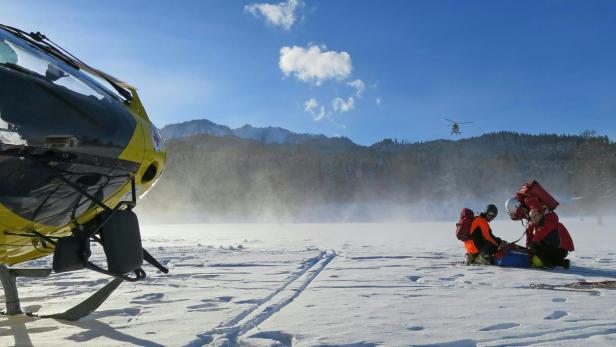 Ein Rettungsteam versorgt eine Person auf einer Schneefläche, im Hintergrund ein Rettungshubschrauber.