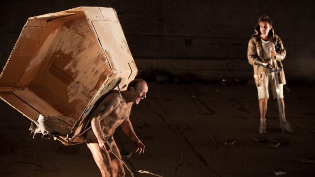 Ein Mann schleppt eine große Kiste auf dem Rücken, während ein anderer mit einem Schwert zusieht.