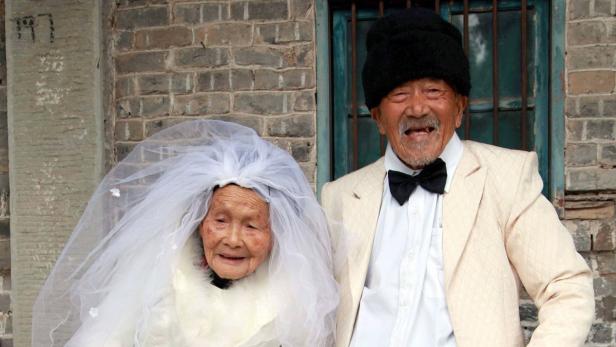 Ein älteres Paar in Hochzeitskleidung posiert vor einer Ziegelmauer.