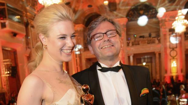 Eine junge Frau und ein Mann im Smoking posieren mit einer goldenen Statue in einem festlichen Saal.