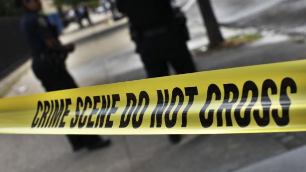 Ein gelbes Absperrband mit der Aufschrift „Crime Scene Do Not Cross“ vor Polizisten.