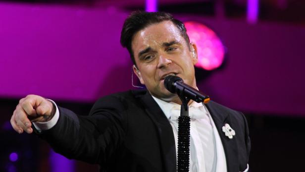 Robbie Williams singt in einem schwarzen Anzug auf einer Bühne.