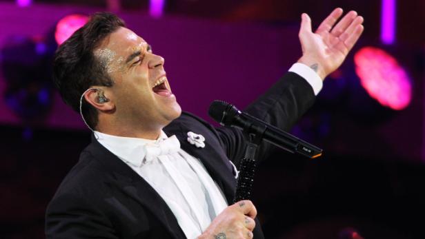 Robbie Williams singt mit erhobener Hand in einem schwarzen Anzug auf der Bühne.
