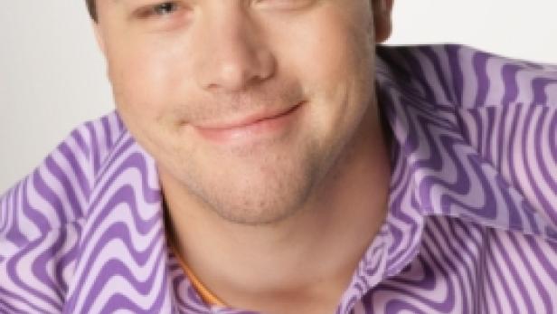 Ein Mann mit blonden Haaren trägt ein lila-weiß gemustertes Hemd und lächelt.