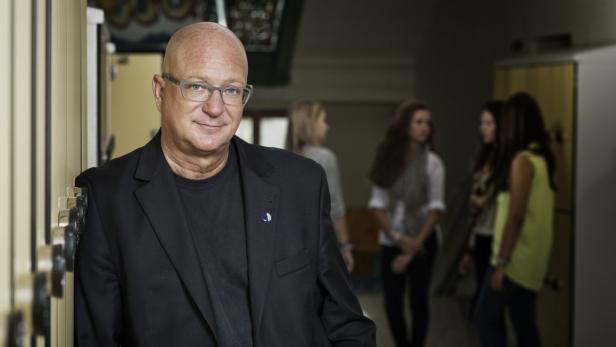 Ein Mann mit Brille lehnt an einer Schließfachanlage, im Hintergrund stehen drei junge Frauen.