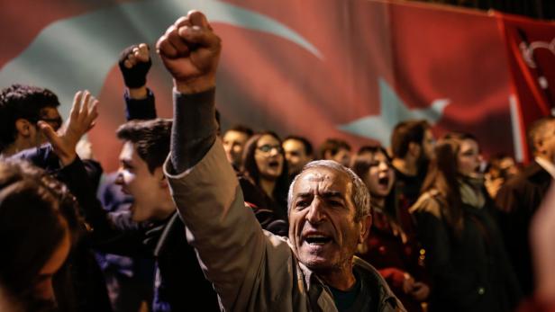 Eine Menschenmenge demonstriert mit erhobenen Fäusten vor einer türkischen Flagge.