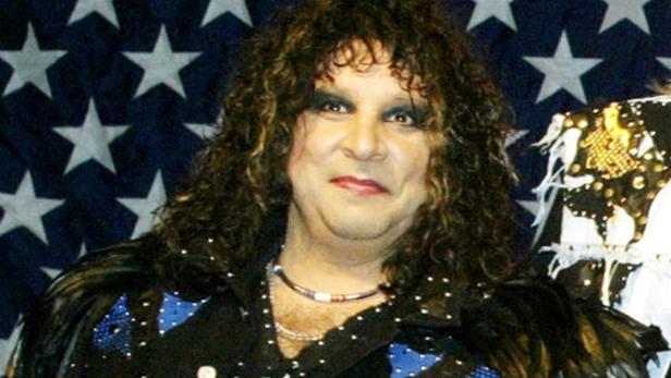 Ein Mann mit dunklen, gelockten Haaren und Make-up vor einer US-Flagge.