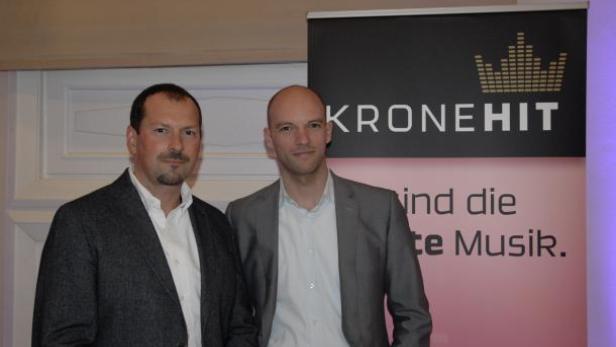 Zwei Männer posieren vor einem „Kronehit“-Banner.
