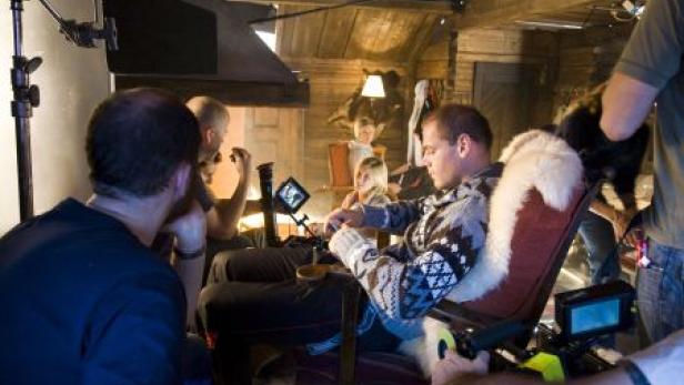 Ein Mann sitzt in einem Sessel auf einem Filmset, umgeben von Filmcrew und Ausrüstung.