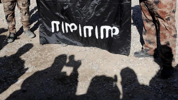 Personen stehen mit einer schwarzen Flagge mit weißer arabischer Schrift auf einem steinigen Untergrund.