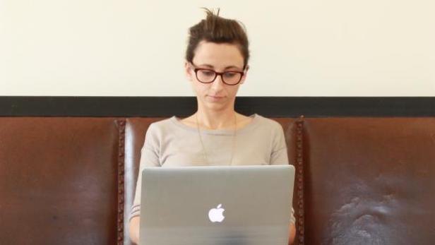 Eine Frau mit Brille sitzt auf einem Sofa und arbeitet an einem Laptop.
