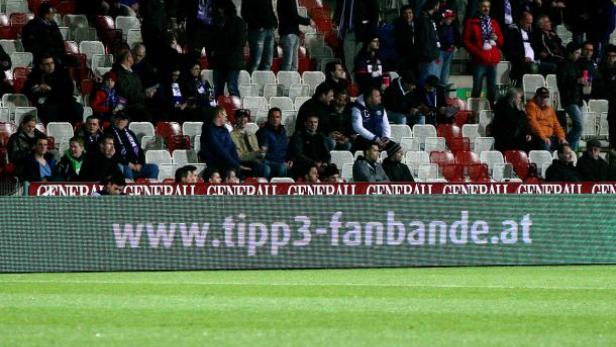 Zuschauer sitzen auf der Tribüne eines Fußballstadions vor einer Werbetafel.