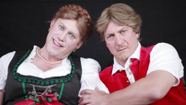 Zwei Männer, einer in Dirndl-Tracht und der andere in Lederhosen-Optik, posieren nebeneinander.