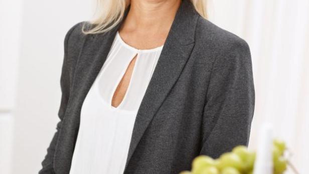 Eine blonde Frau in Blazer und Bluse lächelt in die Kamera. Auf dem Tisch steht eine Etagere mit Obst.