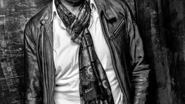 Ein Mann mit Brille, Lederjacke und Schal posiert vor einer dunklen Wand.