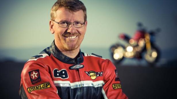 Ein lächelnder Mann in Rennjacke posiert vor einem Motorrad.