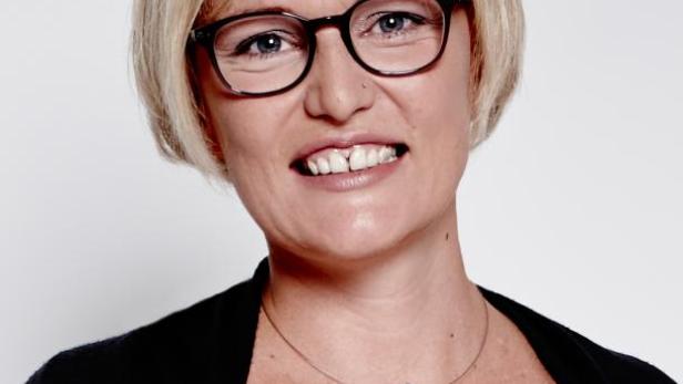 Eine Frau mit blonden Haaren, Brille und einer Halskette mit einem großen Anhänger lächelt.