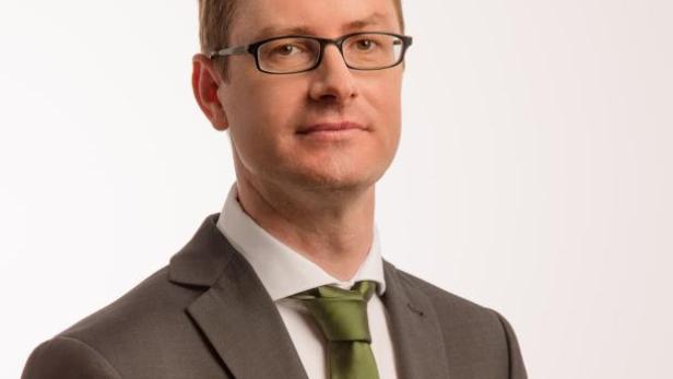 Ein Mann mit Brille, Anzug und grüner Krawatte vor einem weißen Hintergrund.