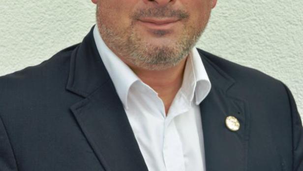 Ein Mann mit Anzug und weißem Hemd vor einer hellen Wand.