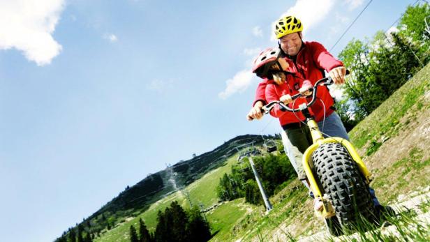 Ein Mann und ein Kind fahren mit einem gelben Mountaincart einen grünen Hügel hinunter.