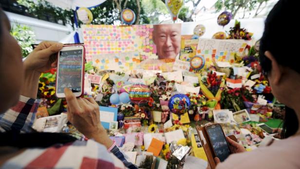 Zwei Personen fotografieren eine Gedenkstätte mit Blumen, Ballons und einem Porträt.