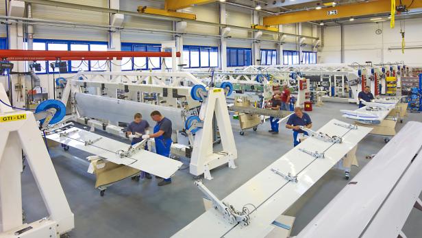 In einer Fabrikhalle arbeiten mehrere Personen an der Montage von Flugzeugflügeln.