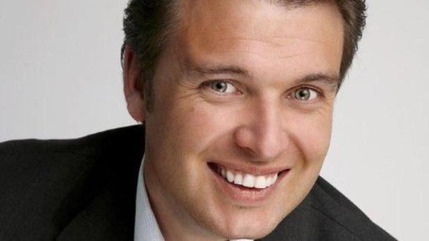 Ein lächelnder Mann in einem dunklen Anzug vor einem hellen Hintergrund.