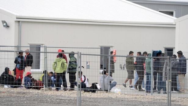 Eine Gruppe von Menschen steht und sitzt vor einem Gebäude hinter einem Zaun.