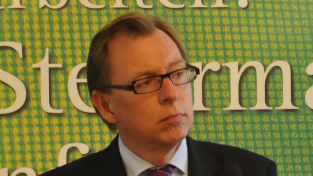 Ein Mann mit Brille und Anzug steht vor einem grünen Hintergrund.