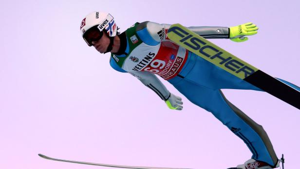 Ein Skispringer in der Luft mit blauer Kleidung und gelben Handschuhen.