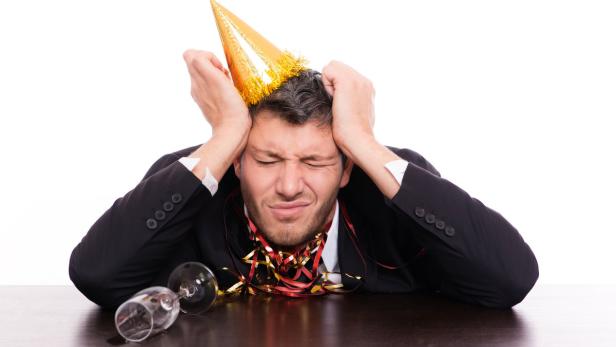 Ein Mann mit Partyhut und Konfetti wirkt verkatert an einem Tisch mit umgekippten Gläsern.