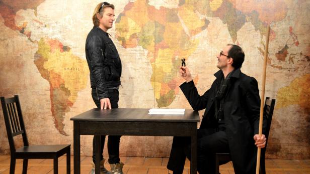 Zwei Männer stehen bzw. sitzen an einem Tisch vor einer Weltkarte.