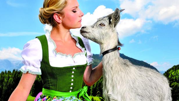 Eine junge Frau in Tracht interagiert mit einer Ziege vor einer Bergkulisse.