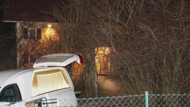 Ein Leichenwagen steht nachts vor einem Haus, das von Bäumen umgeben ist.