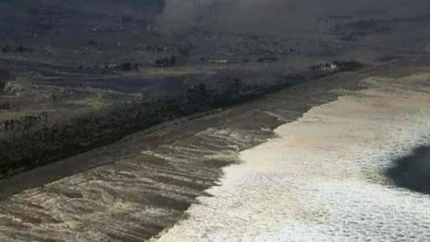 Luftaufnahme einer Küstenlinie mit dunklem Sand und schäumenden Wellen.