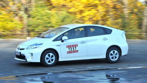 Ein weißer Toyota Prius mit „Kurier Test Kurier“-Aufklebern fährt auf einer Straße.