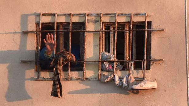 Eine Hand winkt aus einem vergitterten Fenster, an dem Wäsche und ein Schuh hängen.
