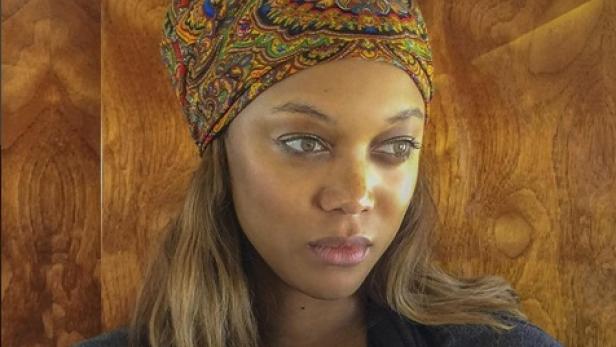 Tyra Banks mit einem bunten Tuch auf dem Kopf vor einer Holzwand.