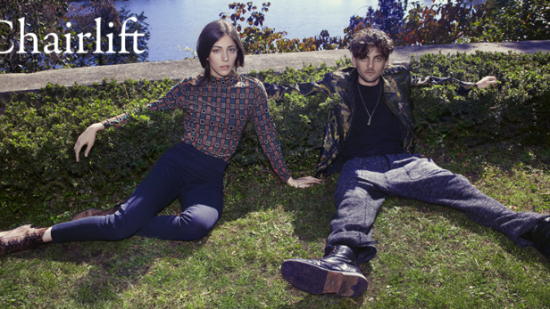 Die Band Chairlift sitzt auf einer Wiese vor einem See.