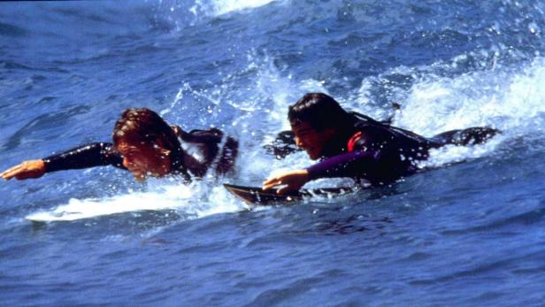 Zwei Surfer paddeln auf ihren Brettern durch die Wellen.